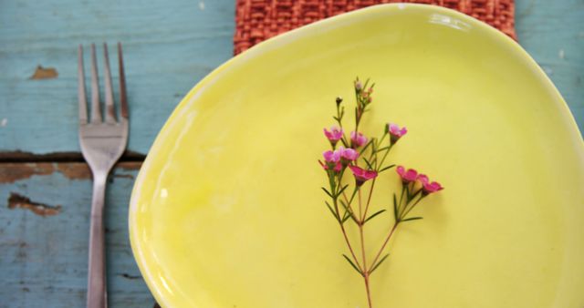 A green plate with a small pink flower on it is placed next to a fork, with copy space. The setting suggests a rustic dining theme with a touch of nature's simplicity.