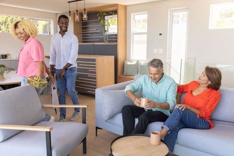 Young couple arriving with gift while senior hosts enjoying mugs in modern living room