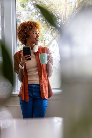 African american woman standing by window holding mug and phone wearing headphones