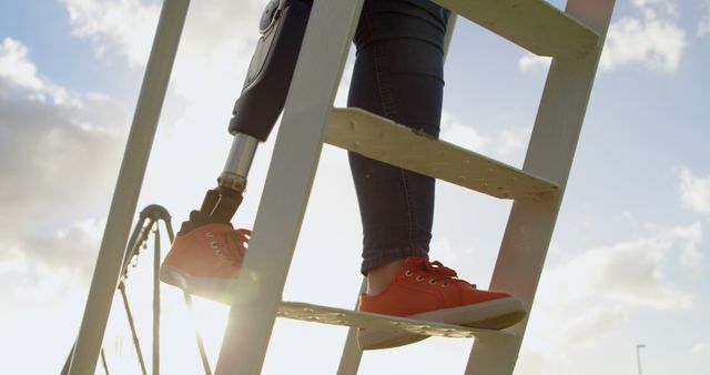 Person with prosthetic leg climbing ladder on a sunny day. Ideal for use in articles or advertising related to mobility aids, resilience, personal strength, outdoor activities, inclusivity, and health products.