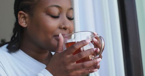African american woman sipping tea by sunlit window with nose hoop and braided hair