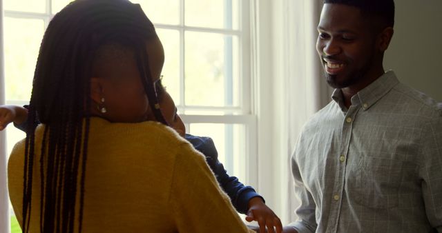 This image shows a family spending quality time together at home. The mother is holding their child while the father smiles lovingly at them. The natural light coming through the window adds warmth to the scene. Ideal for use in advertising campaigns focused on family values, home life, parenting products, or emotional well-being.