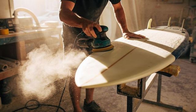 Craftsman sanding surfboard in sunlit workshop showing stringer and three fins