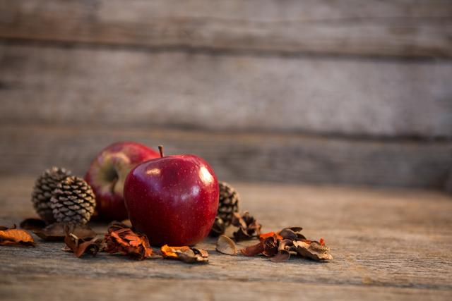 Red apple and pine cones resting on weathered wooden plank surrounded by dried autumn leaves. Ideal for holiday decorations, autumn themes, natural and rustic visuals, and festive décor ideas.