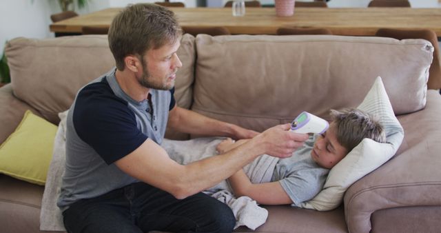 A father measures his sick son's temperature with an infrared thermometer while the boy rests on a couch. Could be used for articles or adverts on family healthcare, parenting tips, and home care for ill children.