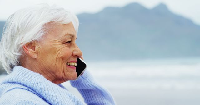 Elderly woman with white hair talking on smartphone while standing by beach. Wearing a blue sweater and smiling, capturing a moment of relaxation and connection. Suitable for use in advertisements, articles on senior lifestyle, technology use among elderly, or health and wellness.