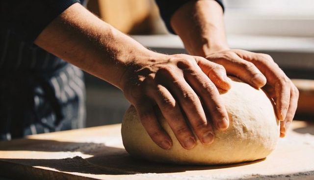 Shaping Hands Molding Artisan Bread Dough on Wooden Board in Sunlit Bakery Workspace