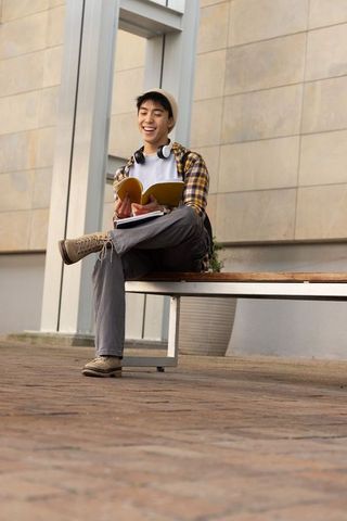 Smiling asian teen studying outdoors on bench with headphones and yellow notebook in urban plaza