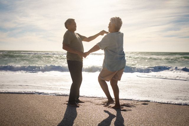 A senior couple joyfully dancing on a beach with waves crashing in the background. Ideal for use in advertisements or articles focusing on retirement, love in later years, healthy living, and active senior lifestyles.