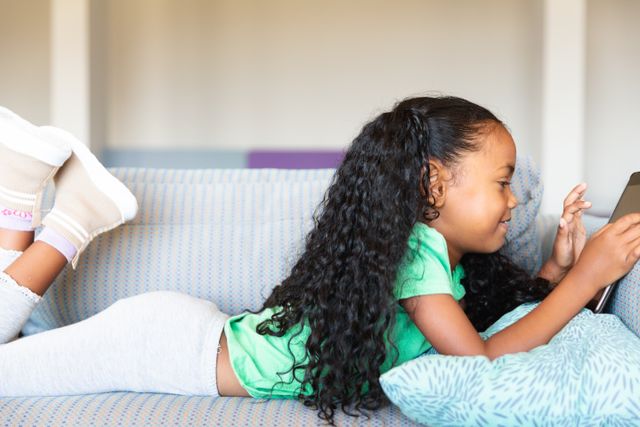 Young African American girl lying on a sofa using a digital tablet. Ideal for educational content, technology use in children, home learning environments, and relaxation themes. Suitable for articles, blogs, and advertisements focusing on childhood education, digital learning tools, and family lifestyle.