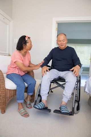 Asian couple sharing caregiving moment, partner supporting man in wheelchair at home