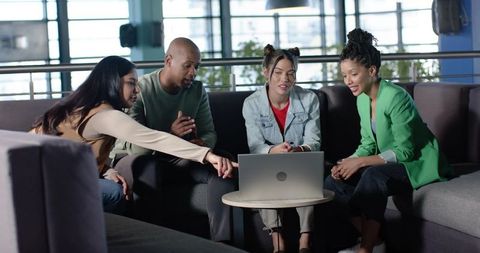 Diverse team collaborating around laptop in modern lounge pointing and brainstorming together