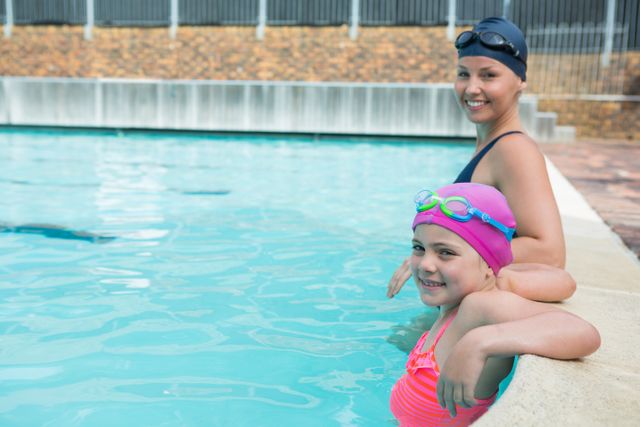 Female instructor and young girl relaxing in pool, both wearing swim caps and goggles. Ideal for use in materials promoting swimming lessons, water safety, fitness, and leisure activities. Perfect for illustrating the bond between instructor and student, and the enjoyment of swimming.