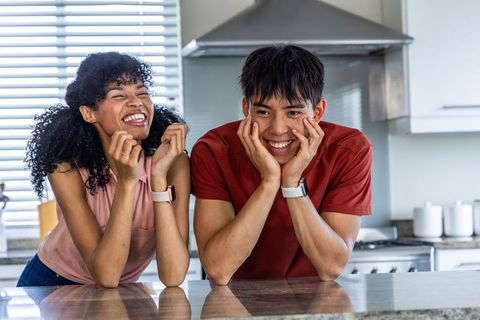 Laughing multicultural couple leaning on countertop wearing smartwatches, modern lifestyle