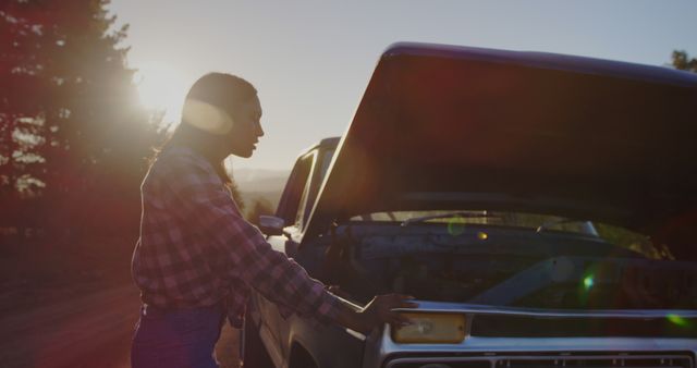 Young African American woman examines car engine outdoor. She troubleshoots a vehicle issue during a sunset road trip.