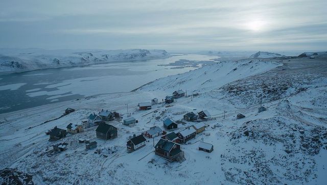 Remote Arctic village perching on snowy hillside overlooking frozen fjord at dawn