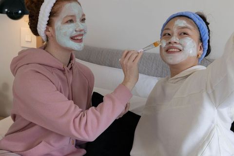 Caucasian and asian friends applying facial masks on bed, enjoying cozy home spa night