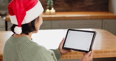 Senior asian woman wearing santa hat holding tablet at cozy kitchen island with candles