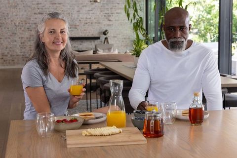 Sunlit diverse senior couple enjoying breakfast with orange juice in modern open-plan kitchen