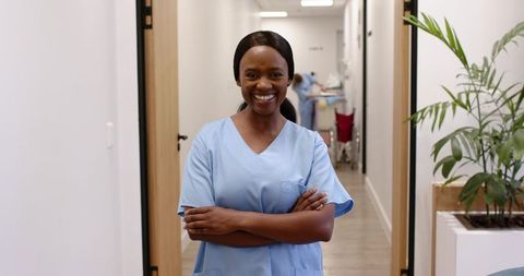 Smiling Black Nurse Standing in Clinic Corridor with Arms Crossed, Confident Healthcare