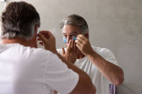 Mature man applying under-eye gel patches while looking into bathroom mirror