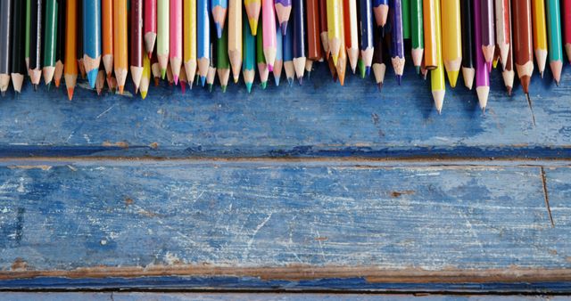 A vibrant array of colored pencils is lined up against a textured blue wooden background, with copy space. The assortment of pencils suggests creativity, art, and the potential for drawing or coloring.