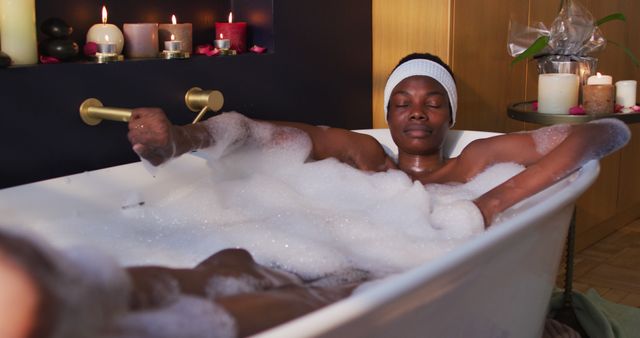 African American woman taking a relaxing bubble bath. Scene includes lit candles providing a serene ambiance. Appropriate for wellness blogs, spa services advertisements, and self-care promotional material.