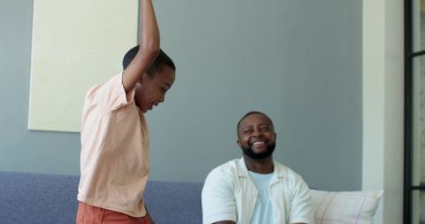African american father smiling while son raising arms on sofa in cozy modern living room
