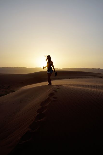 Woman walking on sand dunes during sunset, creating a striking silhouette. Perfect for use in travel blogs, adventure magazines, exploration-themed advertising, or nature documentaries. Highlights themes of solitude, beauty of nature, and wanderlust.