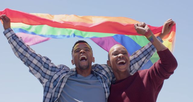 Happy african american gay male couple celebrating with pride flag outdoors in the sun. Togetherness, relationship, pride, lgbtq, equality, rights and celebration, unaltered.