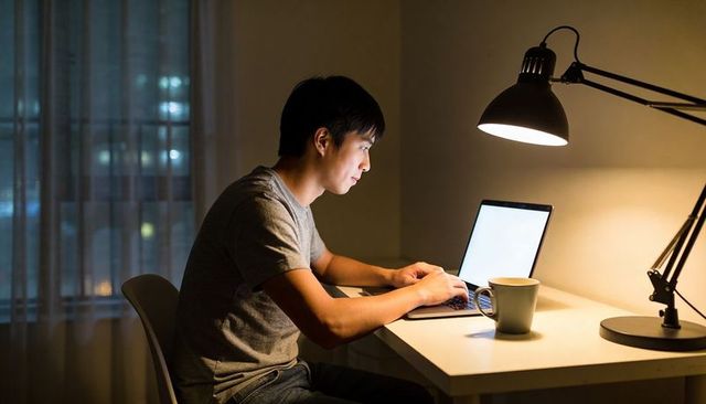 Asian young man working late on laptop at home desk with lamp and coffee cup at night