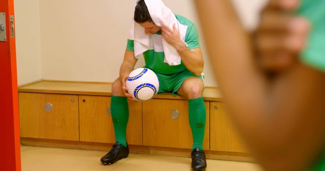 Soccer player taking a break in locker room, showing signs of exhaustion by holding towel over head and ball on lap. Great for illustrating post-game moments, the physical demands of sports, athletes recovering, or promoting sports equipment and apparel.