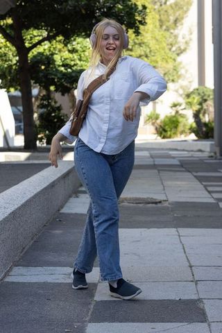 Young woman dancing while walking with pink headphones and brown crossbody bag in city