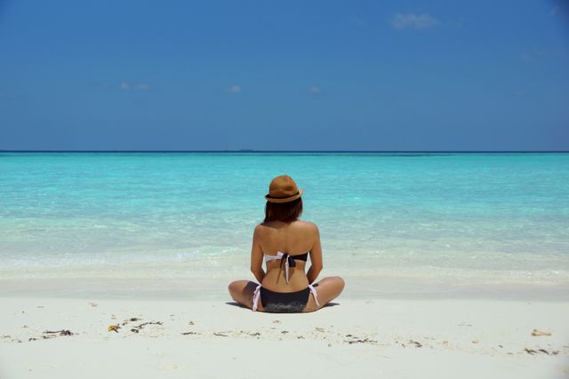 This image shows a woman sitting on a tropical beach, facing the turquoise water, and wearing a hat and swimwear. Ideal for travel blogs, vacation brochures, relaxation and wellness promotions, beachwear advertisements, and social media posts focused on summer or beach themes.