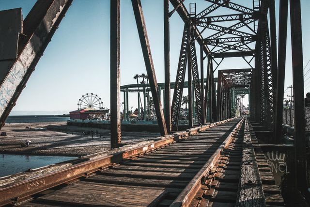 Old railway bridge made of steel leading across river to an amusement park visible in background with ferris wheel and coastal attractions. Ideal for illustrating themes of industrial heritage, tourism, travel adventure, and seaside entertainment.