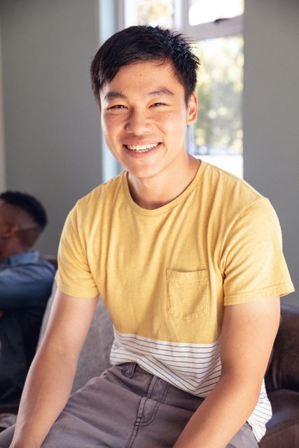 Young man sitting on a sofa at home, smiling and looking at the camera. He is wearing a casual yellow shirt and appears relaxed and happy. This image can be used for lifestyle blogs, home living articles, or advertisements promoting a cheerful and relaxed home environment.