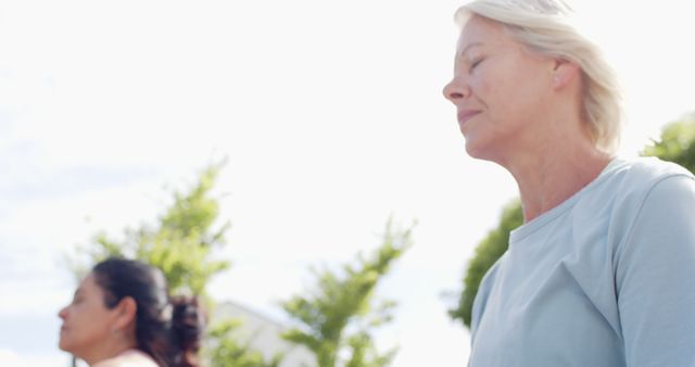 Two senior women meditating outdoors in a sunlit setting, eyes closed, appearing calm and serene. This image can be used for promoting wellness, mindfulness, healthy living, and outdoor activities. Ideal for wellness programs, health articles, and lifestyle campaigns.