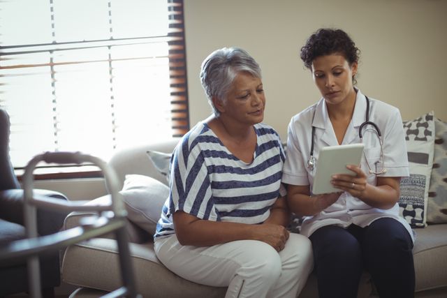 Female doctor discussing over digital tablet with senior woman in living room at home