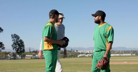 Coach giving guidance to baseball players in green uniforms on sunny field during practice