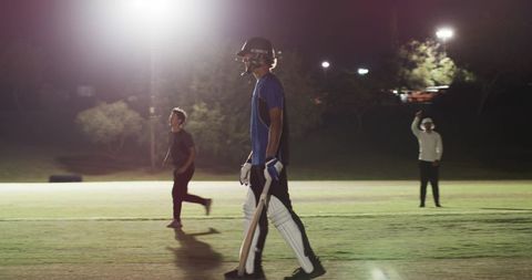 Night cricket batsman walking with helmet and bat under floodlights casting long shadows