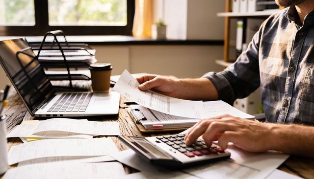 Calculating bearded man in plaid shirt reviewing bills with laptop and calculator