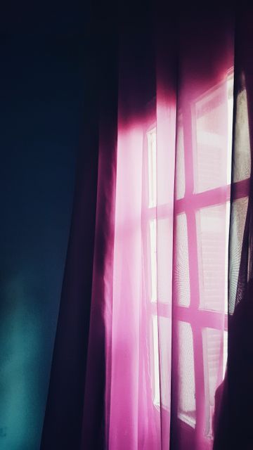 Magenta curtains gently sway while sunlight streams through a window, creating a contrast with the dark surroundings. This image can be used for themes of solitude, tranquility, introspection, or design inspirations for home interiors.