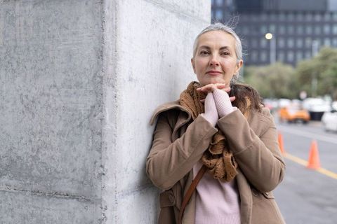 Mature woman leaning on concrete column clasping hands wearing coat scarf on urban sidewalk