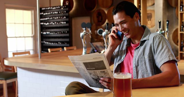 Young man sitting at brewery bar, reading newspaper and smiling while talking on the phone. Holds a pint of beer in front of him. Suitable for themes of leisure, communication, modern lifestyle, and relaxation in a casual setting.