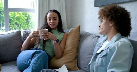 African American friends chatting on cozy gray sofa, drinking coffee and sharing notes