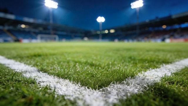 Floodlit football pitch corner closeup showing marked white turf line and goal in distance
