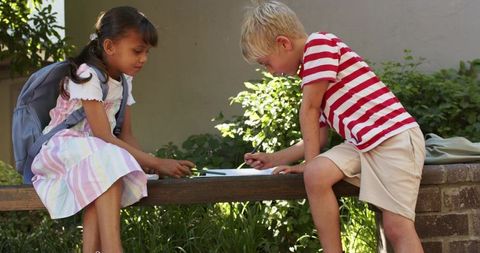 Young classmates sharing notebook on sunlit schoolyard bench studying together