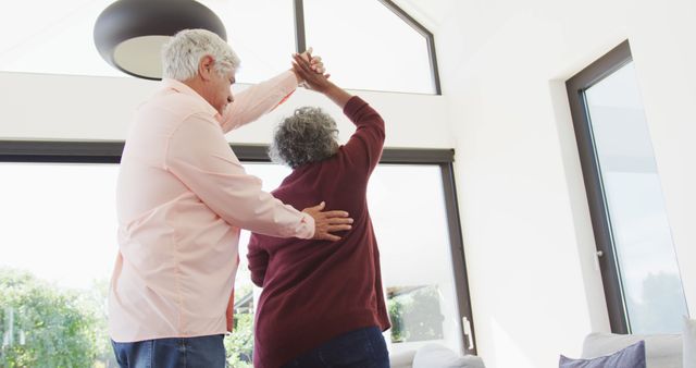 Elderly couple dancing in modern living room with large windows, creating bright and cheerful atmosphere. Useful for projects involving family, love, aging, and active lifestyle. Good for senior living advertisements, health and wellness promotion, and celebrating togetherness.