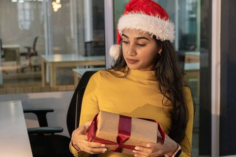 Woman wearing santa hat inspecting wrapped gift in modern open-plan office at night