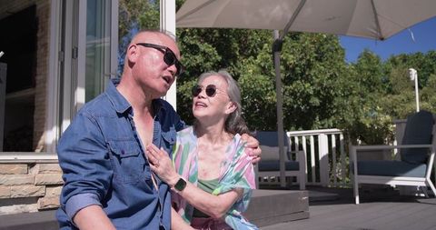 Asian senior couple relaxing on patio wearing sunglasses and smartwatch, colorful summer
